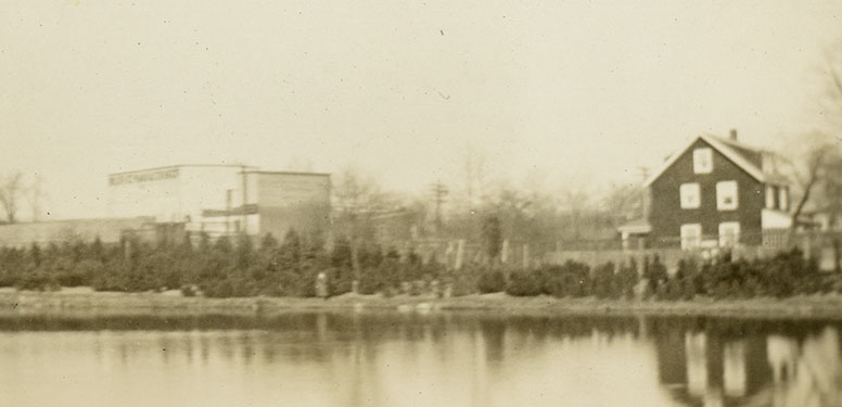 Pond with three-story shingled home and warehouse building in background.