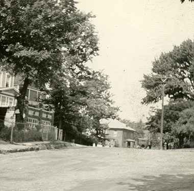 Nurse standing at bottom walkway of a house on a curving road with trees and other houses.