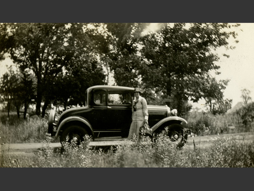 White female nurse in uniform standing beside a Ford Model A Coupe on a rural road with trees.