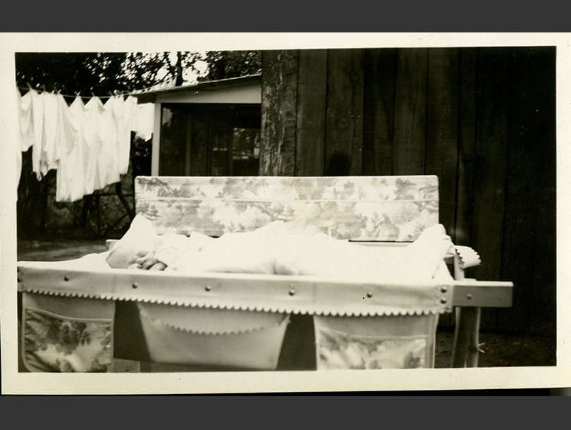 Swaddled infant rests on a wash table in a backyard, laundry hanging in the background.