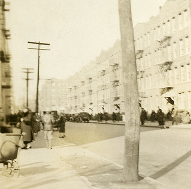 City street with four-story building on right, electricity poles on left, and pedestrians.