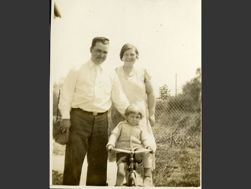 Mother and father standing posed behind a toddler boy on his tricycle.