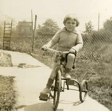 Toddler boy in sweater and shorts riding a tricycle on a cement walkway.