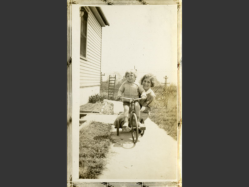 Toddler boy posing on his tricycle on a walkway, with a White nurse in her uniform kneeling behind.