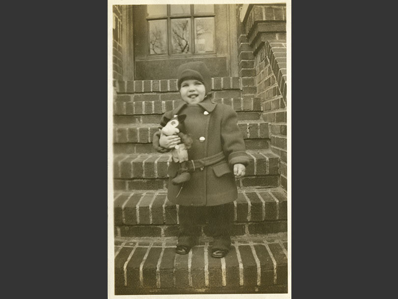 Holding a toy, a toddler boy in coat and hat stands on brick front steps.