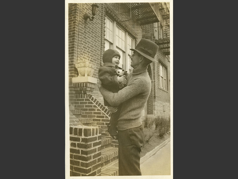 Next to brick steps, a father in hat holds his toddler son, who has a toy in his hands.