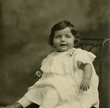 One-year-old girl in a dress sitting on a metal scrollwork chair.