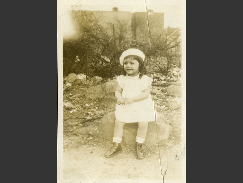 Toddler girl, wearing a dress and beret, sitting on a rock.