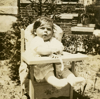 An 8-month girl sitting outside in a small wooden chair that has a little attached shelf in front.
