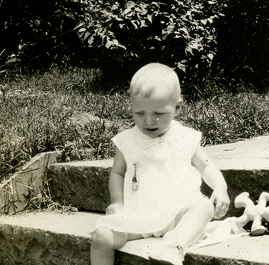 A 10-month White girl sitting on steps to a walkway, a toy beside her.
