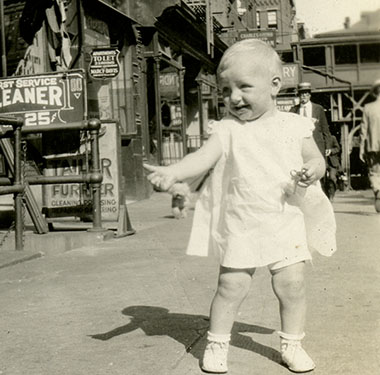 An 11-month White girl standing on a street in 1930s New York, signs for businesses in background.
