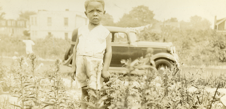 African American toddler boy in shorts standing in a field, 1930s Ford and houses in the background.
