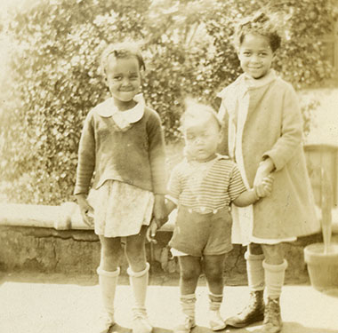 Two African American girls standing next to an African American toddler boy on an outside terrace.
