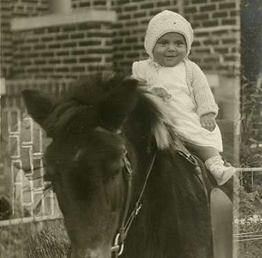 Baby in a crocheted cap sitting on a saddled pony.