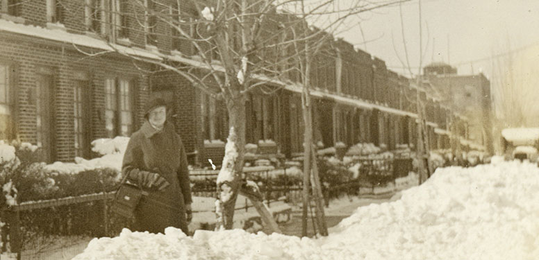 A White female nurse standing in the snow in front of row homes in the city.