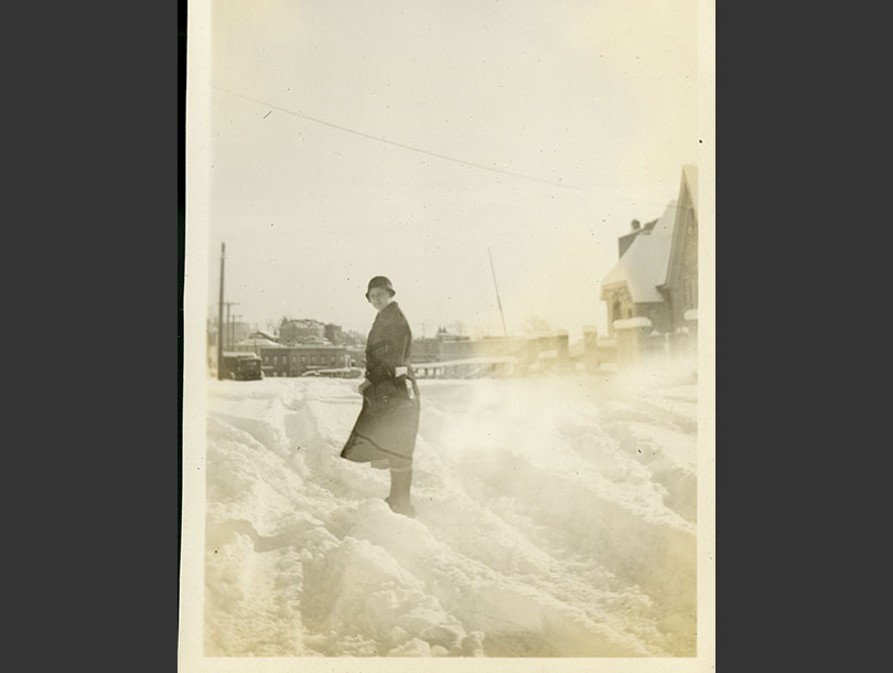 Nurse in coat and cloche hat standing in foot-high drifts of snow, buildings in distance.