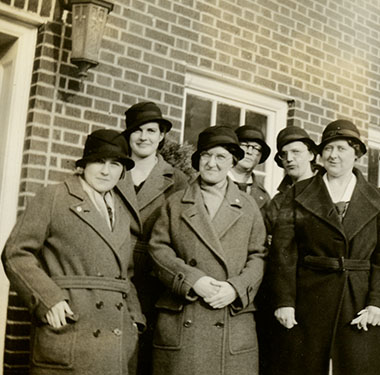 8 smiling, White female nurses in overcoats and cloche hats outside a brick building.