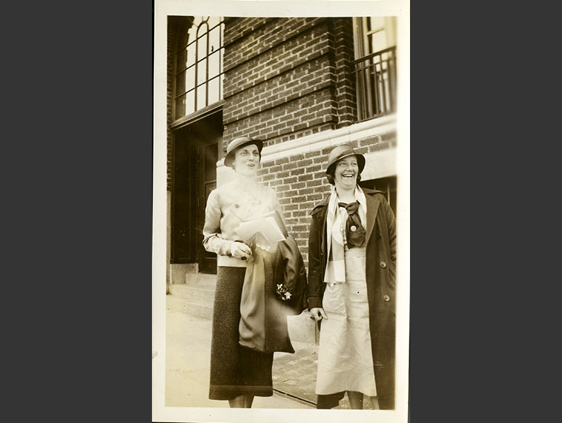 Two, White females in cloche hats standing near a brick building. Miss Giles is in uniform.