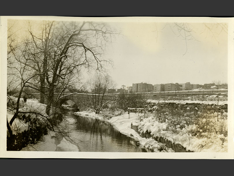 Small stone bridge arching over snowy banks of a narrow stream, apartments in far distance.