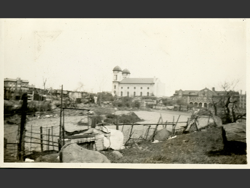 Two-towered church rising in distance beyond a lot jumbled with shacks and various fences.