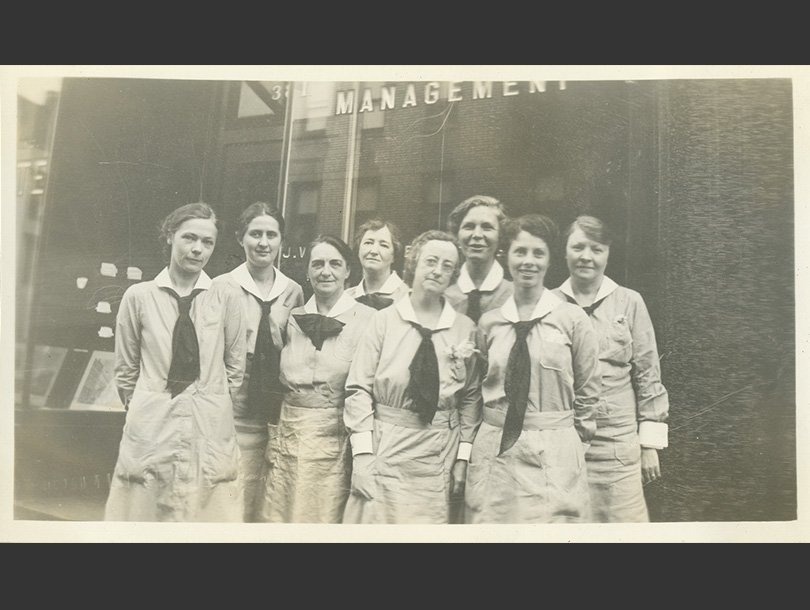 8 White, female nurses in uniform dresses with double front pockets and neck ties in front of a building.