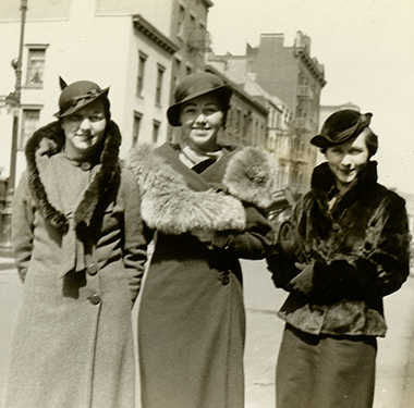 3 White female nursing students in fur-trimmed coats and cloche hats standing on a Manhattan street.