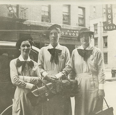 3 White female nursing students in uniform dresses and hats standing on a Manhattan street corner.