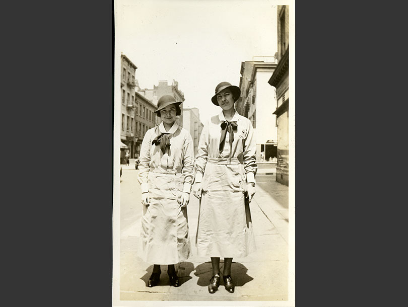 Two female nursing students in uniform dresses and cloche hats posing on a Manhattan sidewalk.
