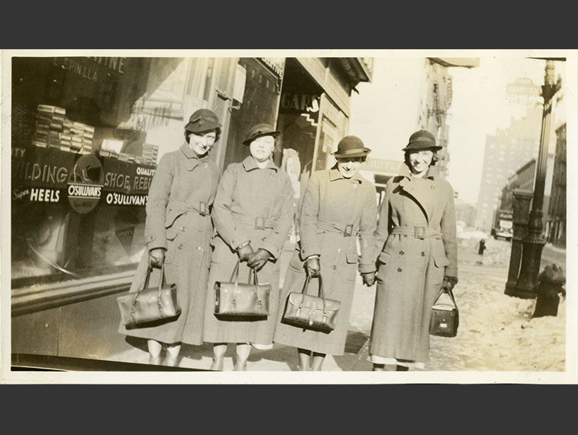 Four White, female nurses in matching winter coats, hats, and nurses’ bags, standing in front of a store.