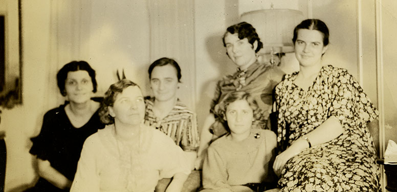 6 women smiling and sitting closely together in a living room.