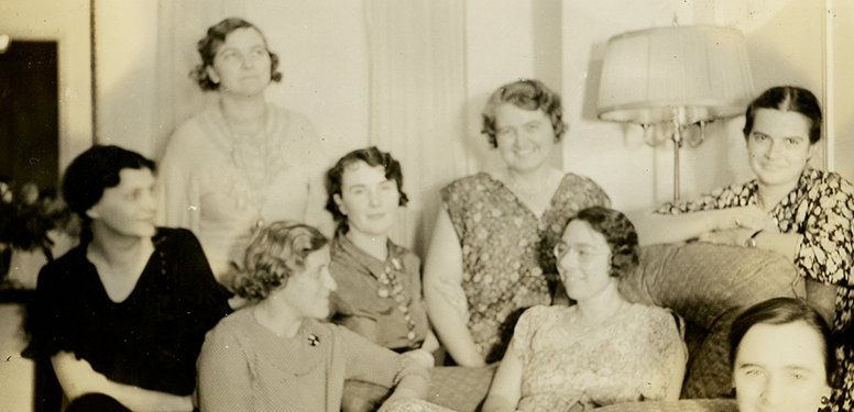 8 women smiling and sitting closely together in a living room.