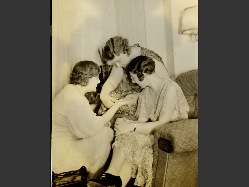 Three women sitting together in a living room, one reading the palm of another.