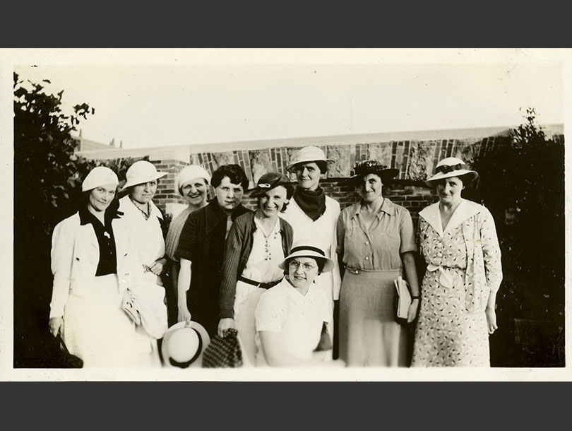 Group portrait of nine White women dressed up and standing near a wall.