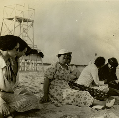 Seven women in dresses sitting on Jones Beach, life guard stand and obelisk water tower in background.