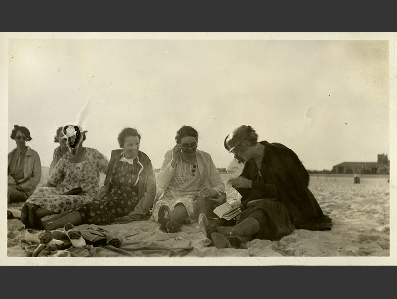Seven smiling women in dresses sitting on Jones Beach, some are laughing while looking at a booklet.