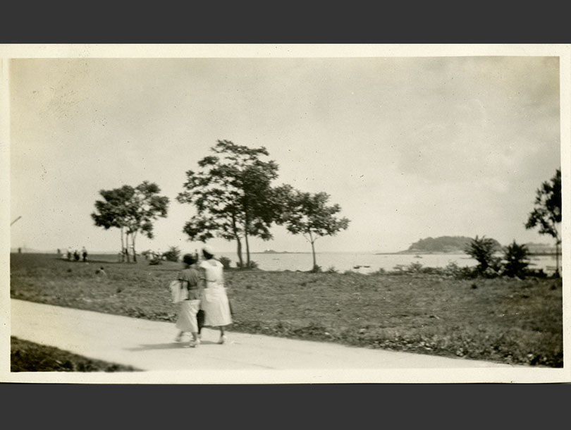 Two women in heeled-shoes walking along a paved path at the seashore.