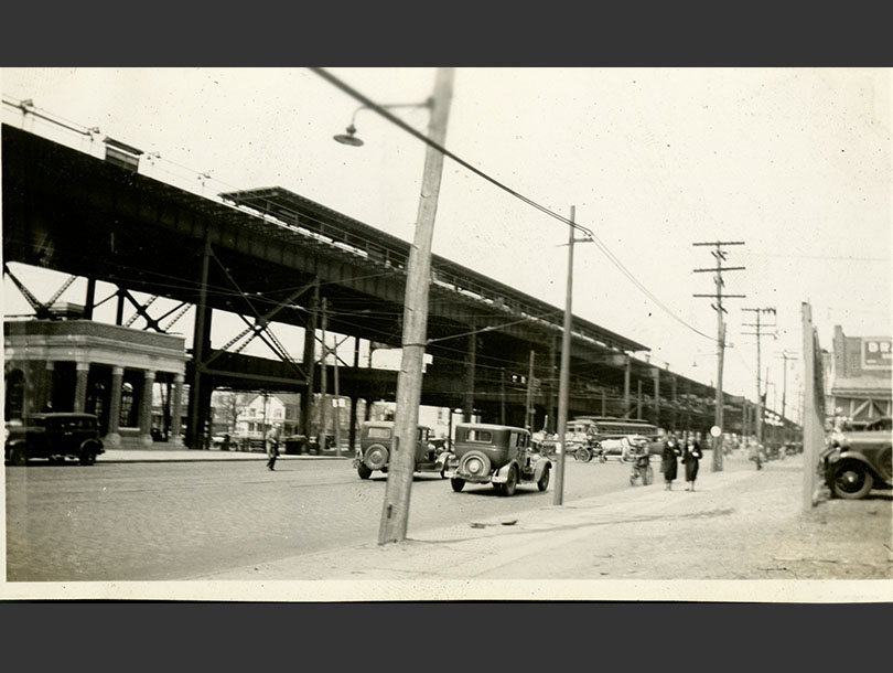 City street showing electricity poles, early 1930s cars, and an elevated train line.