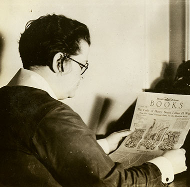Profile view of a White, woman reading the <em>Herald Tribune<em> at a desk, nearby a cat and telephone.