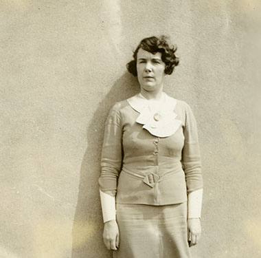 A White woman in a two-piece dress with white shawl collar standing full-length against a wall.