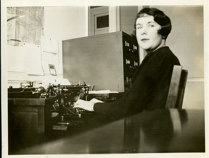 A White woman seated at desk, using a manual typewriter. Filing cabinet in background.