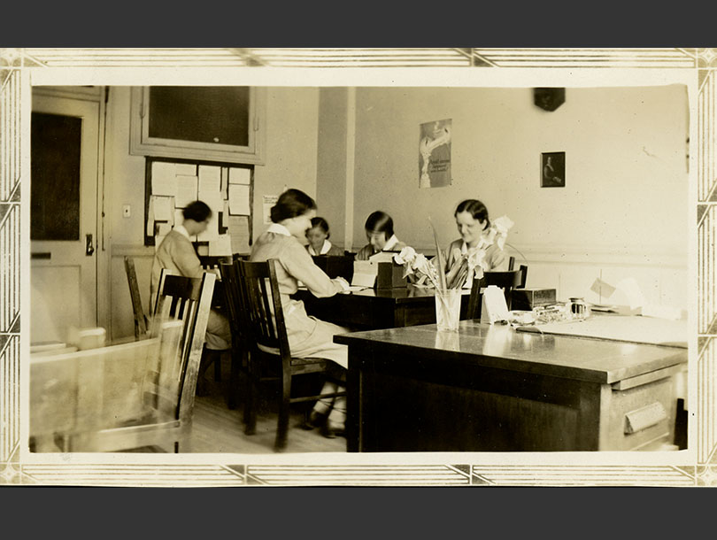 Five female nurses in uniforms working at desks in an office. Bulletin board hanging on the back wall.