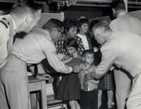 Four uniformed men inspecting two Cuban children’s arms for vaccination marks. Three women appear in background.