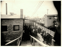 Mexicans of mixed ages wearing hats waiting in a line between a house and a bridge.
