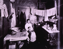 Woman sitting with a child in a cluttered room with laundry hanging from the ceiling and kitchenware scattered.
