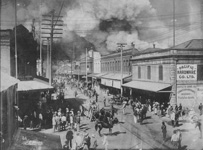 Street view of buildings on fire with people standing in the street and horse drawn carriages.