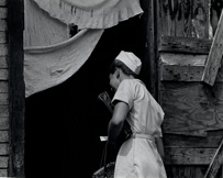 A white, female, uniformed nurse walking into a run-down wooden structure or home.