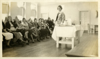 A uniformed, white, female nurse standing next to a table with an audience of seated women in front of her.