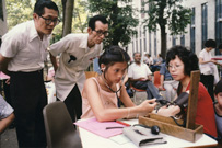 Young Asian woman seated at a table outdoors conducting a blood pressure test on an Asian woman with two Asian men standing behind her.