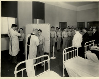 Asian men with bare chests lined up, waiting for inspection by two male medical professionals in white gowns. 