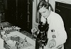 White man in lab coat holds a specimen in front of a microscope.